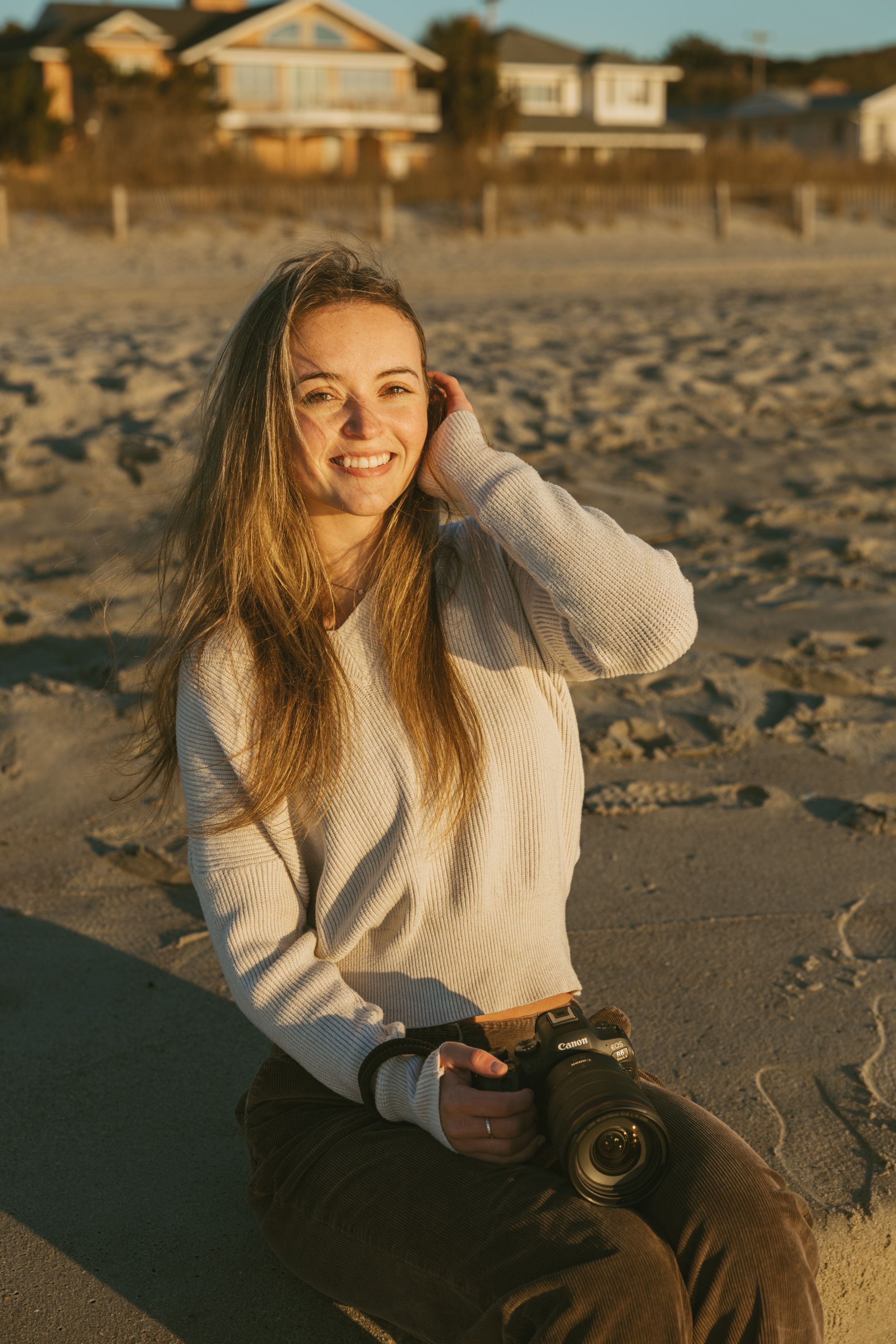 A person in a beige sweater sits on a sandy beach at sunset holding a camera.