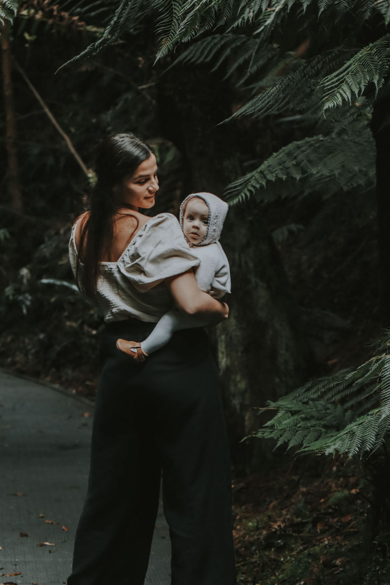 A mother holds her baby in a lush forest setting, both dressed in neutral tones.