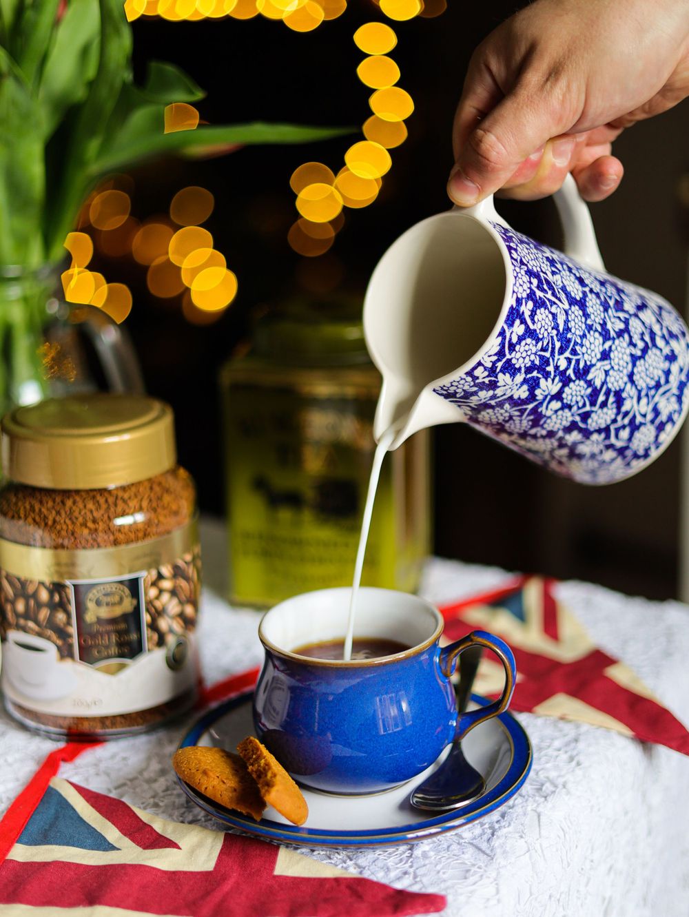 Pouring milk from blue and white patterned pitcher into teacup with bokeh lighting.