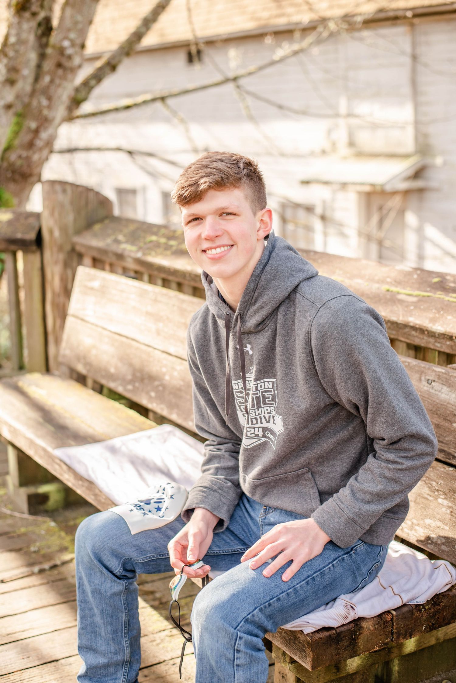 Person in grey hoodie sitting on wooden bench with book in rustic setting.