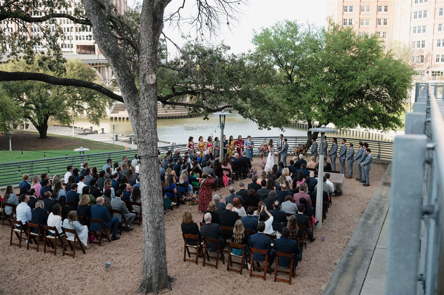 Aerial view of an outdoor wedding ceremony with guests seated beneath large trees.