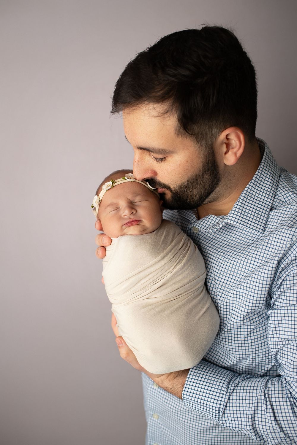 Bearded father tenderly kisses swaddled sleeping newborn baby girl in studio portrait.