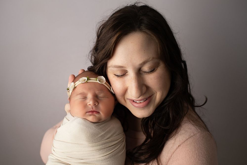 Mother smiles lovingly while holding her swaddled newborn baby girl in studio portrait.