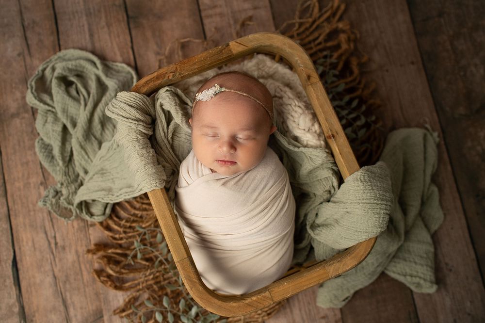 Swaddled newborn baby girl sleeping in a rustic wooden basket with green wraps.