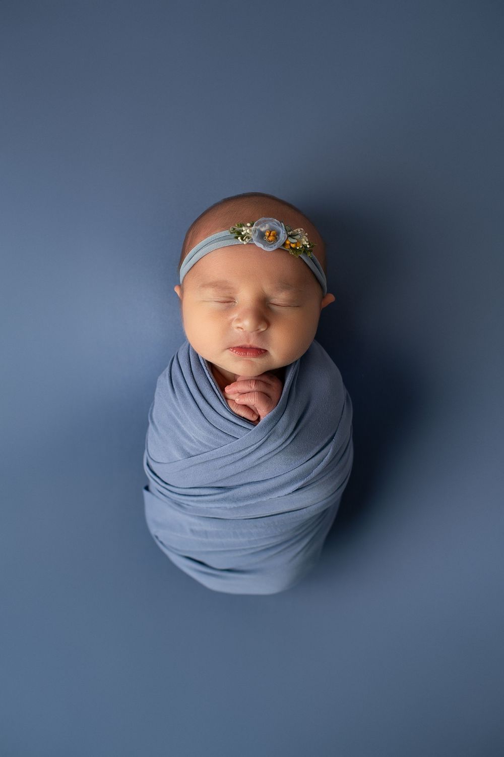 Swaddled newborn baby girl with floral headband lying on a blue background.