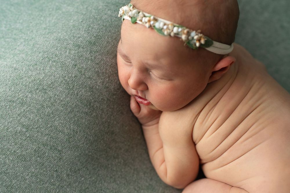 Sleeping newborn baby girl wearing a delicate floral headband on a soft teal backdrop.