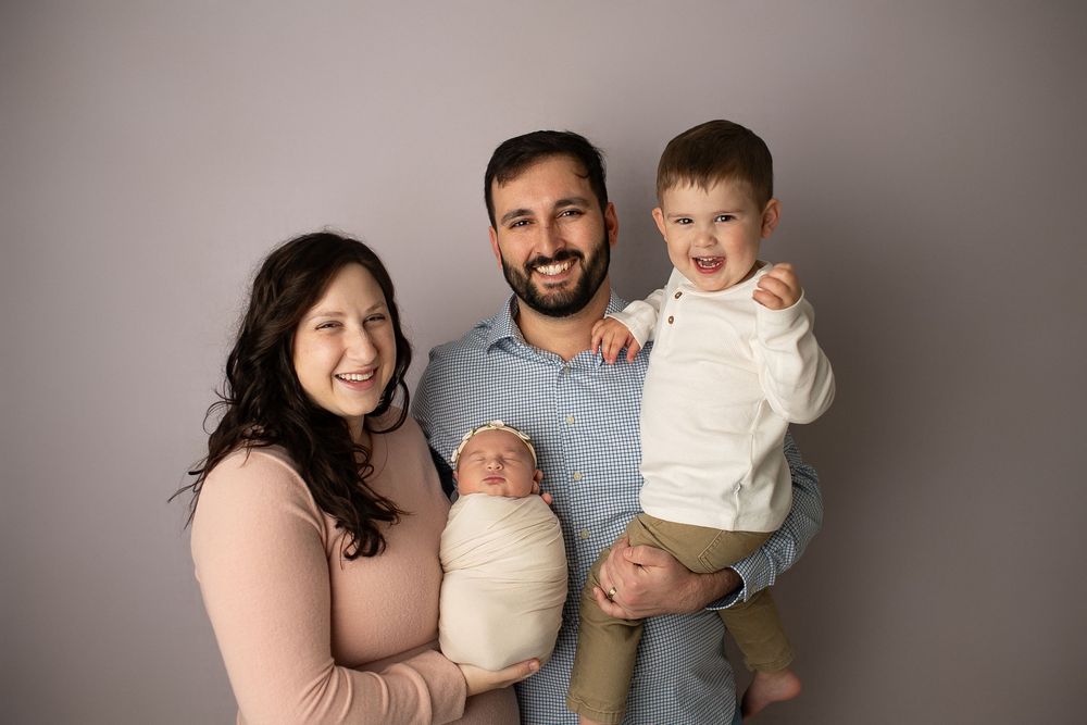 Happy family portrait with parents holding newborn and toddler son against gray backdrop.