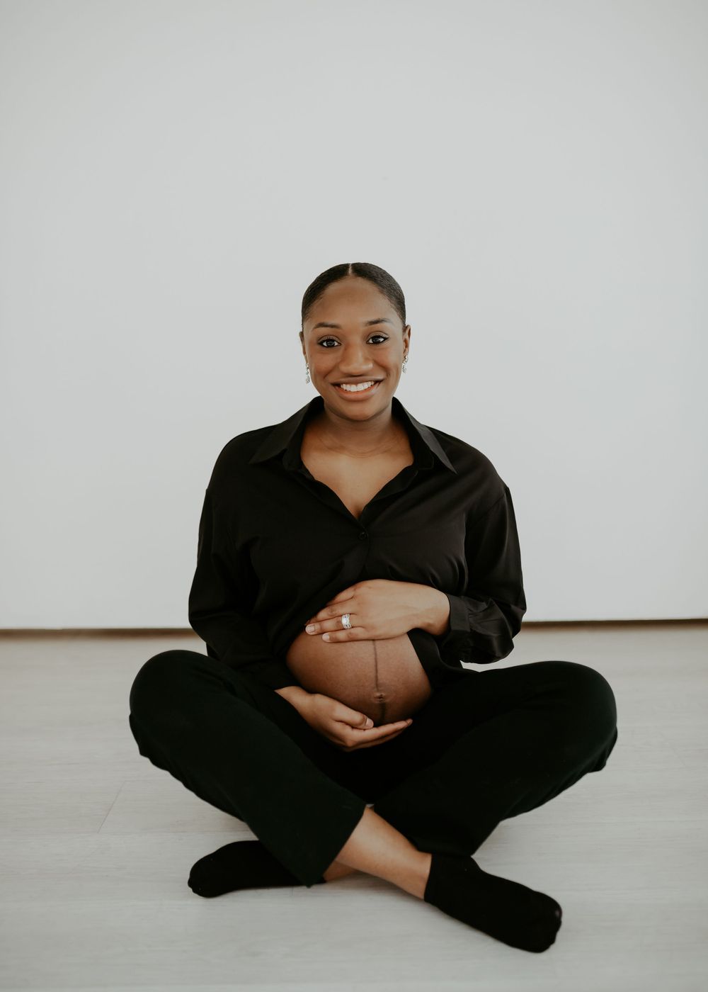 Person in black clothing sits cross-legged on white floor while holding her belly during an in studio maternity session.