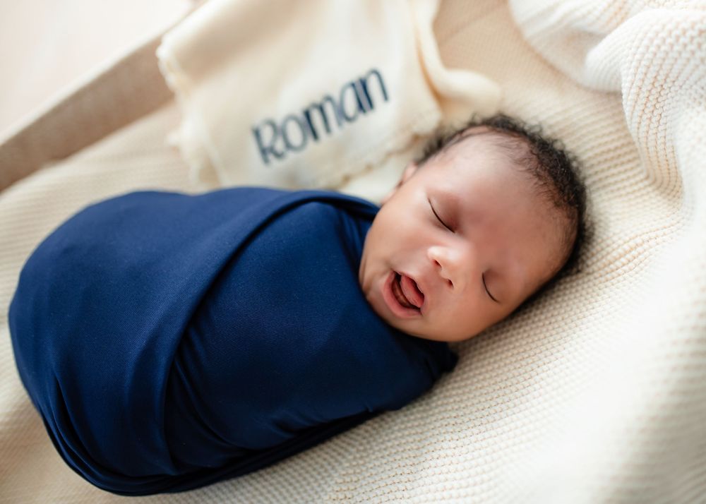 A newborn baby wearing a navy blue sleeper lies peacefully sleeping on white textured bedding during an in studio photo session.