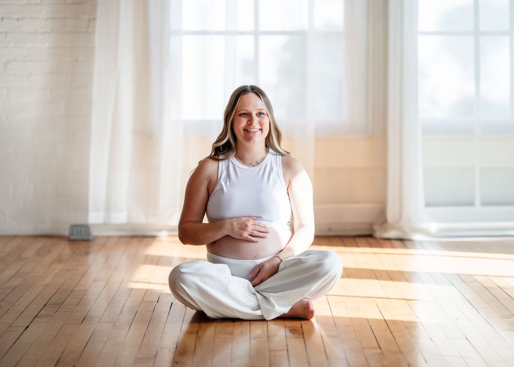 A person in white clothing sits in a lotus position on a sunlit wooden floor near bright windows during a Maternity Session.