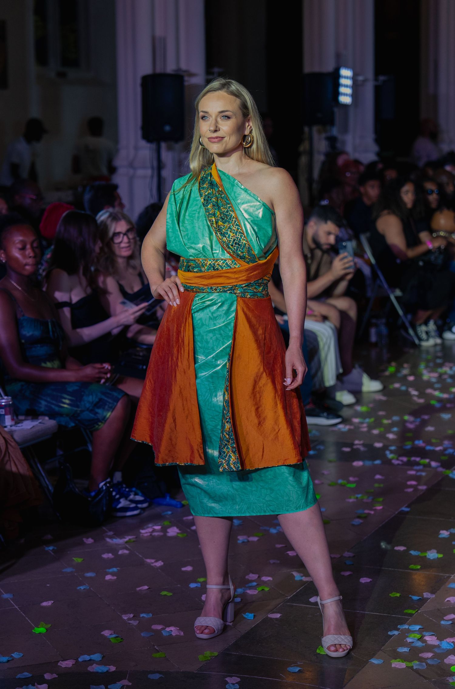 A model walks the runway in a green dress with orange vest during a fashion show with confetti on the floor.