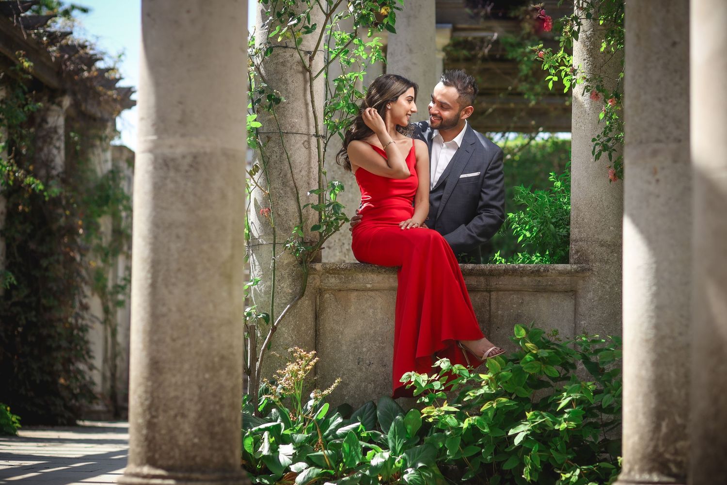 A couple in formal attire shares a romantic moment on a stone ledge surrounded by garden columns and greenery.