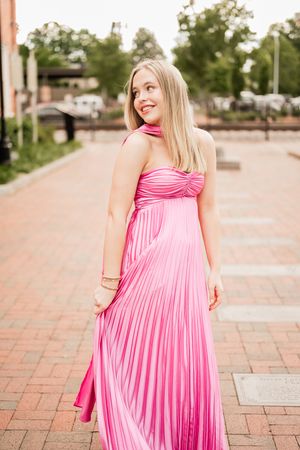 Person in a flowing pink pleated maxi dress twirls on brick pavement in an outdoor setting.