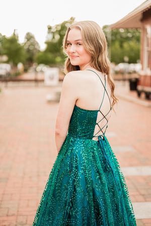 Sparkling emerald green formal gown with sequins and criss-cross back straps shown in outdoor brick setting at sunset.