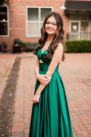 A person wearing a vibrant emerald green formal dress poses on brick pavement in a natural light setting.
