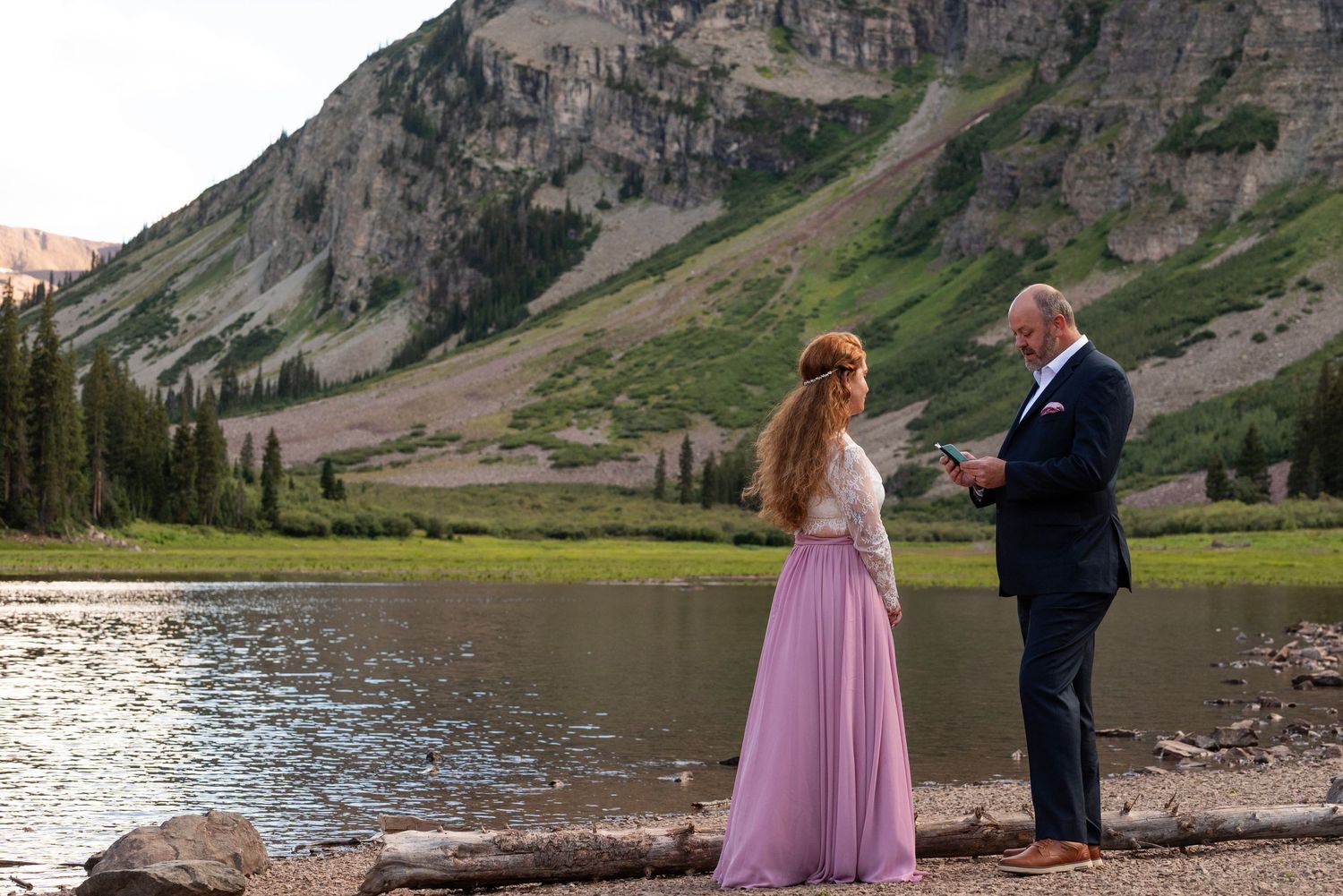 Couple standing by mountain lake with dramatic cliffs in the background.