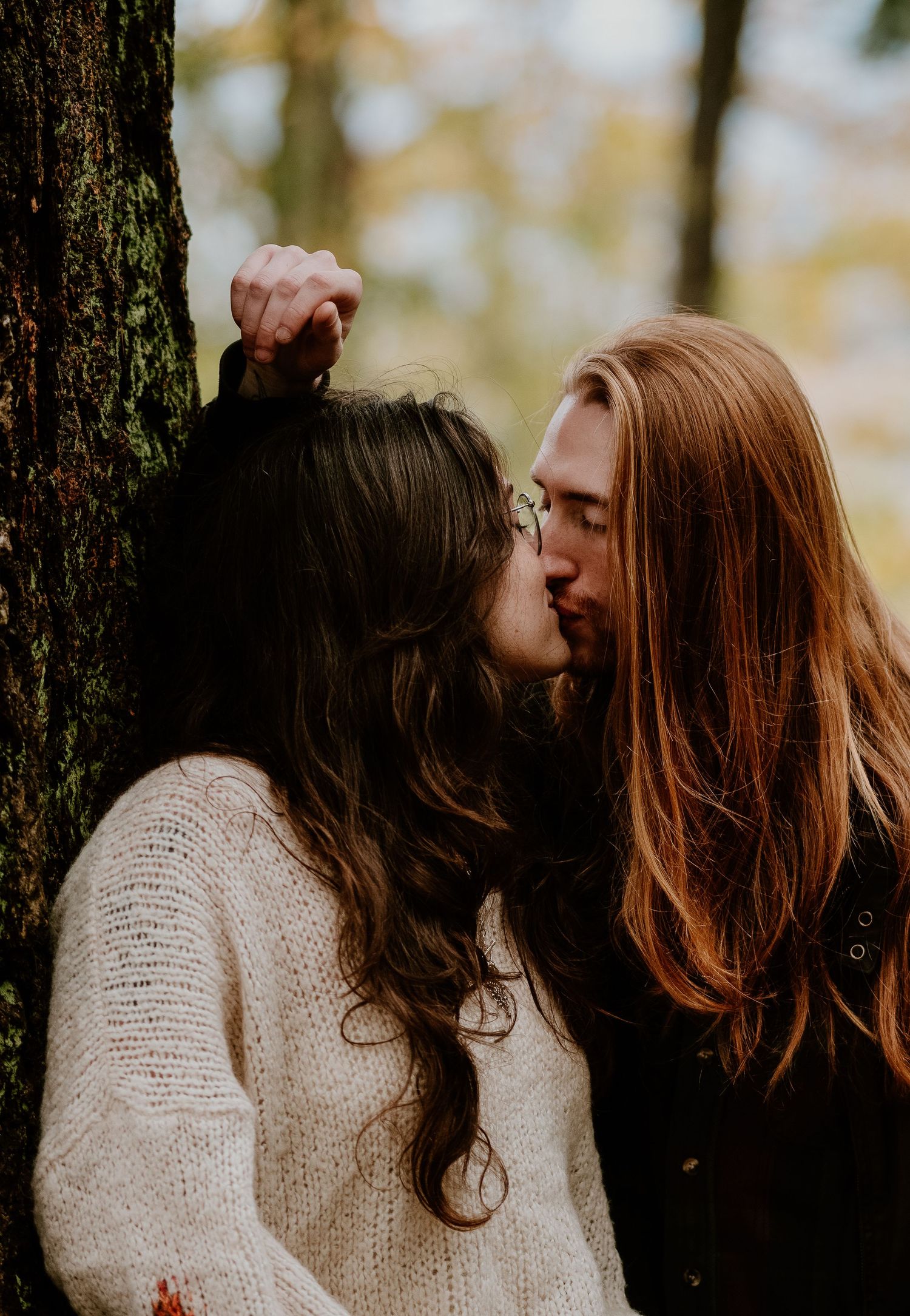 Intimate moment captured between two people wearing cream sweaters against a forest backdrop.