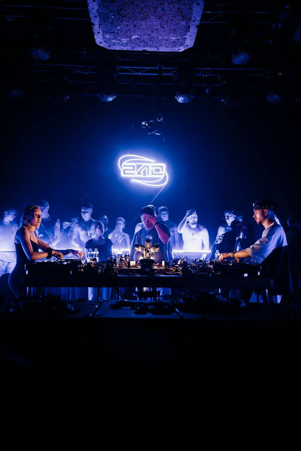 DJs perform at mixing consoles under a neon sign with crowd in background.