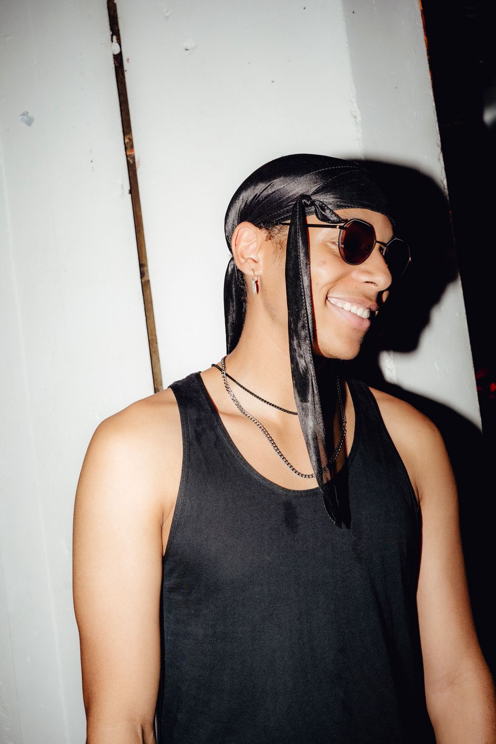 Person in black tank top and sunglasses posing against white wall in artistic photo series.