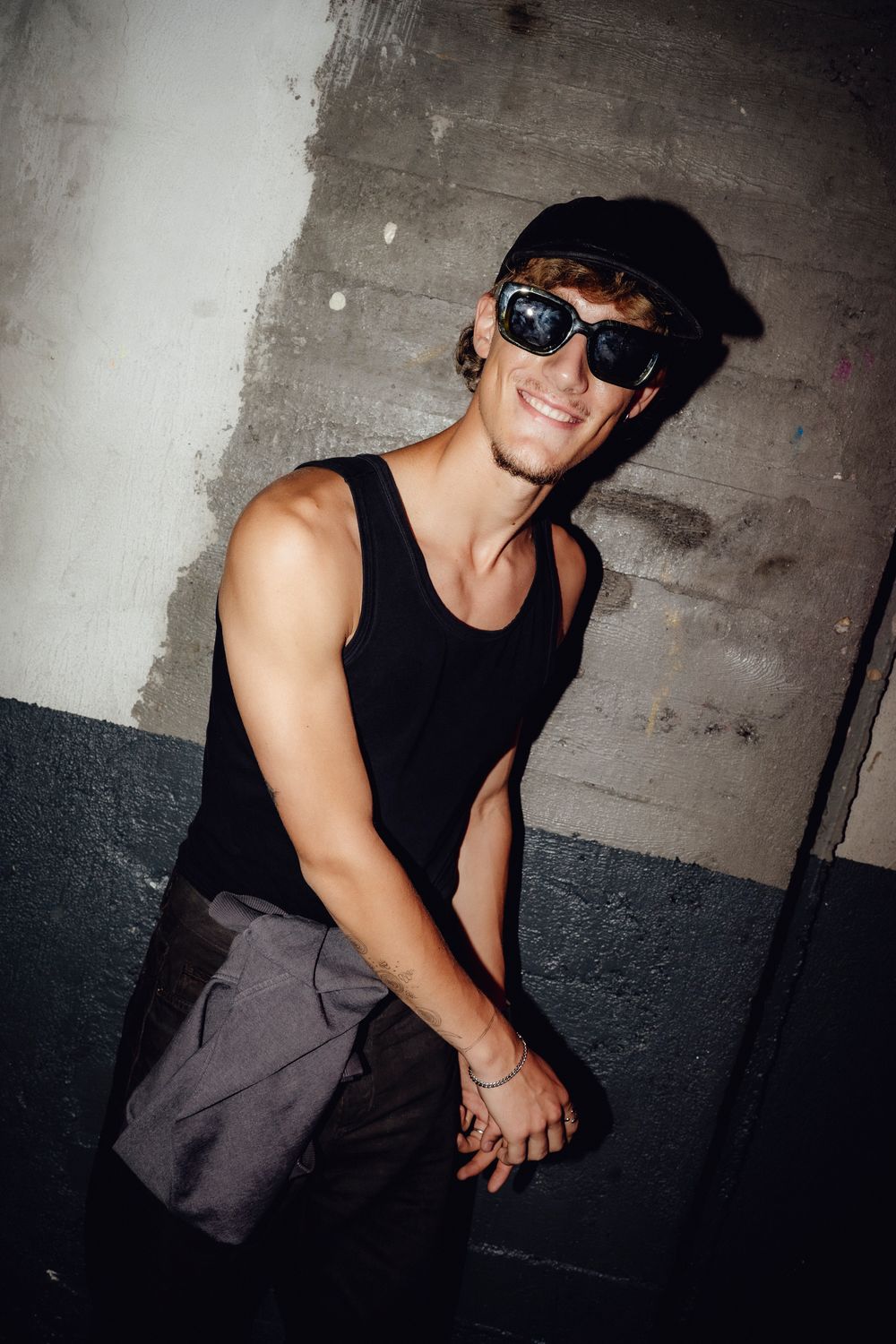 Figure wearing sunglasses and black tank top poses against concrete wall in moody lighting.