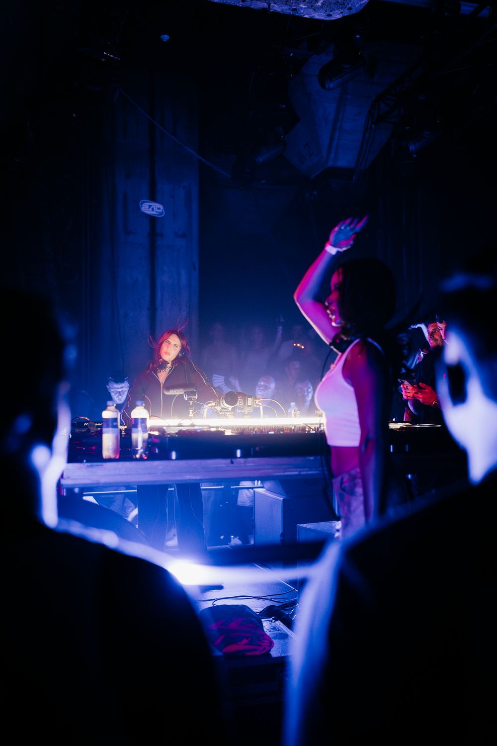 DJ setup illuminated by blue stage lights with performers and crowd silhouettes.