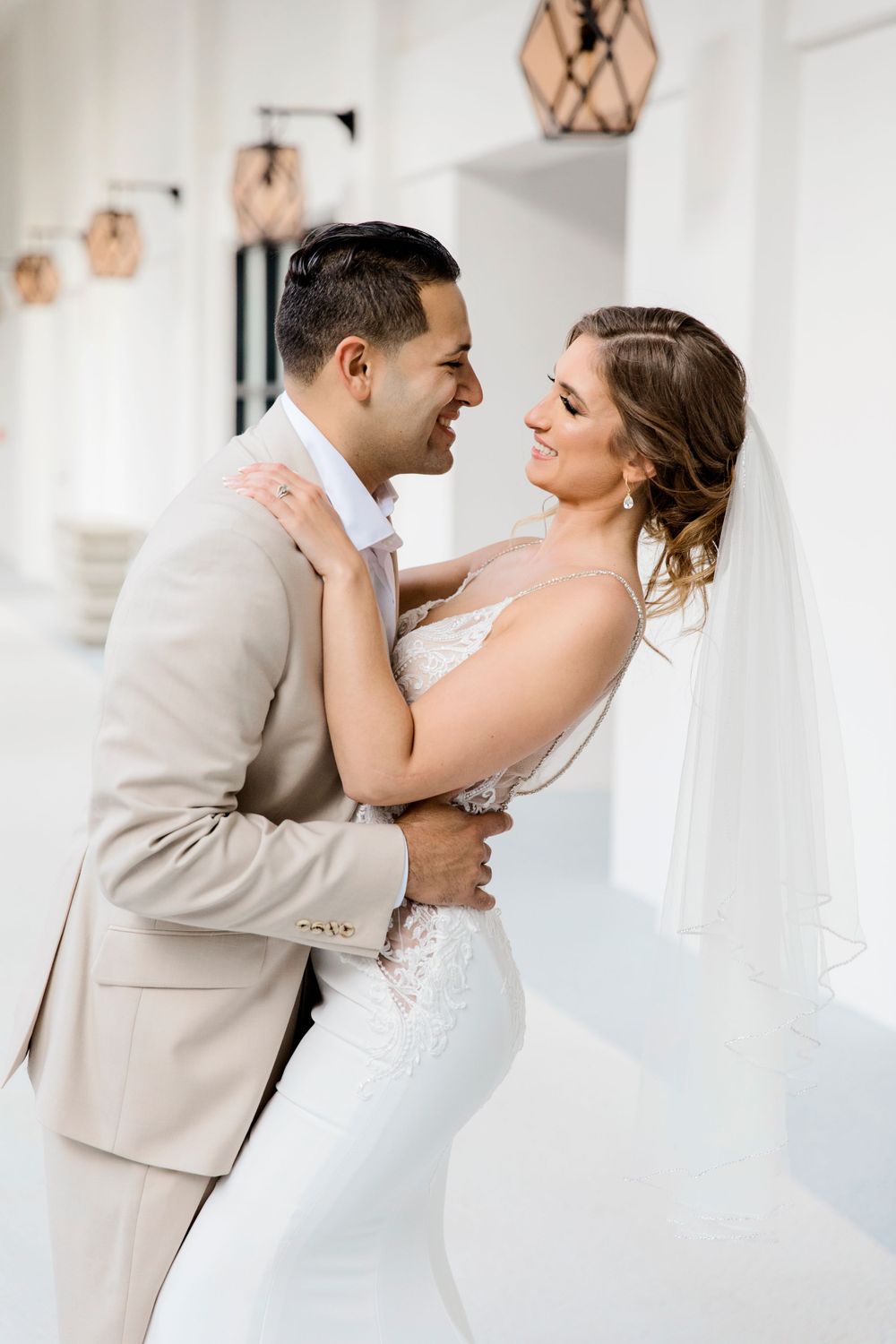 Intimate wedding portrait of couple embracing in elegant white interior with pendant lights.
