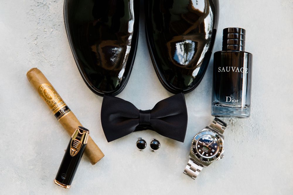 Elegant groom's accessories including patent shoes, bow tie, watch and cologne bottle arranged on white surface.