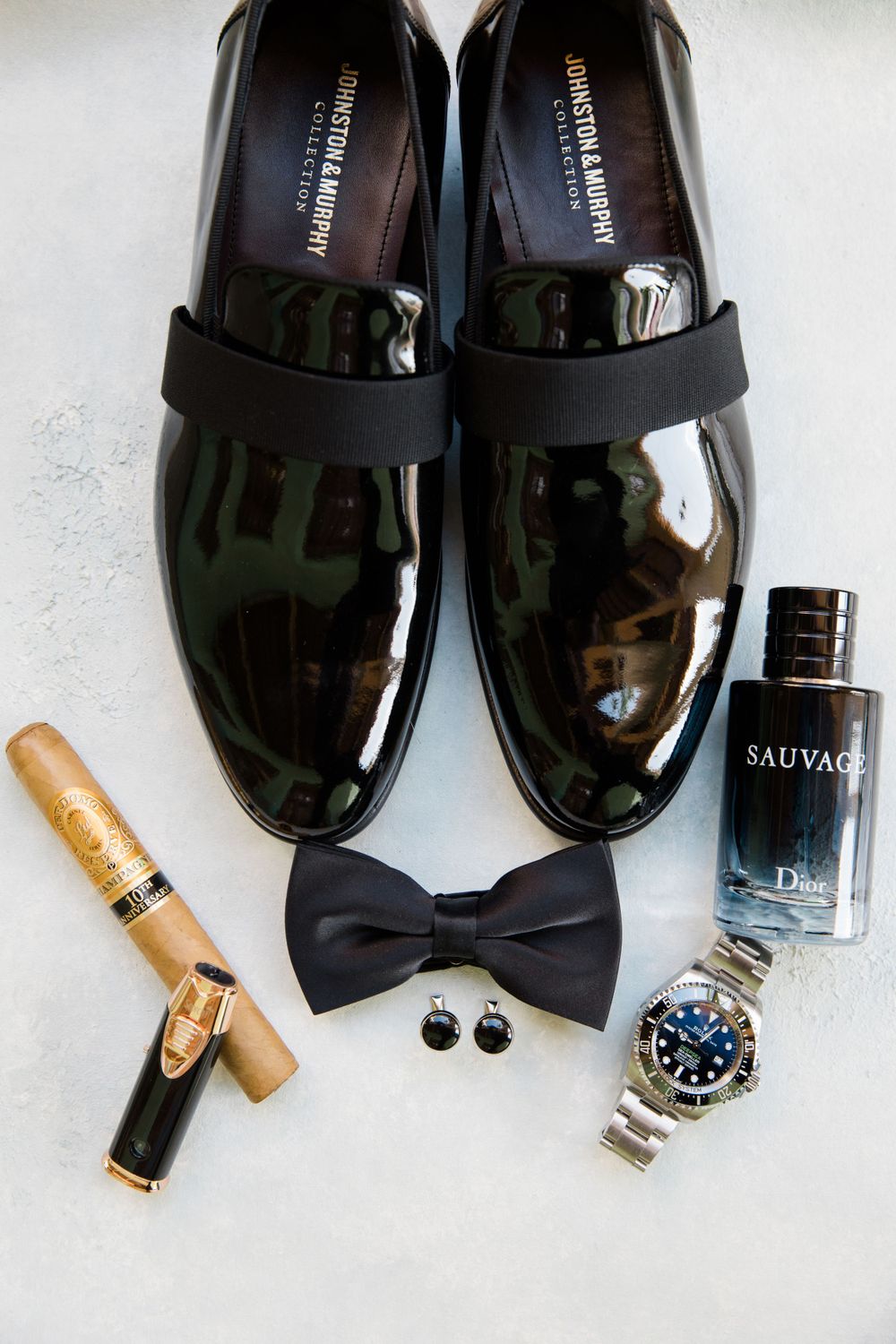 Groom's accessories with patent leather shoes, bow tie, watch, cologne and cufflinks on white surface.