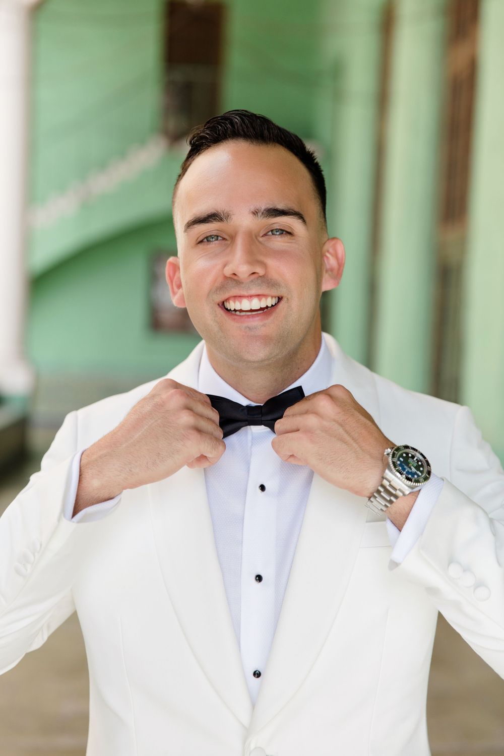 Wedding day portrait in white tuxedo and bow tie against green colonial architecture.