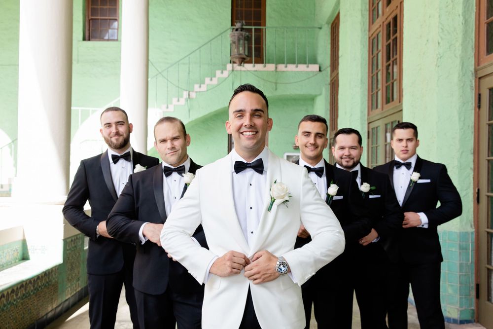 Wedding party in formal attire with groomsmen in black and groom in white tuxedo.