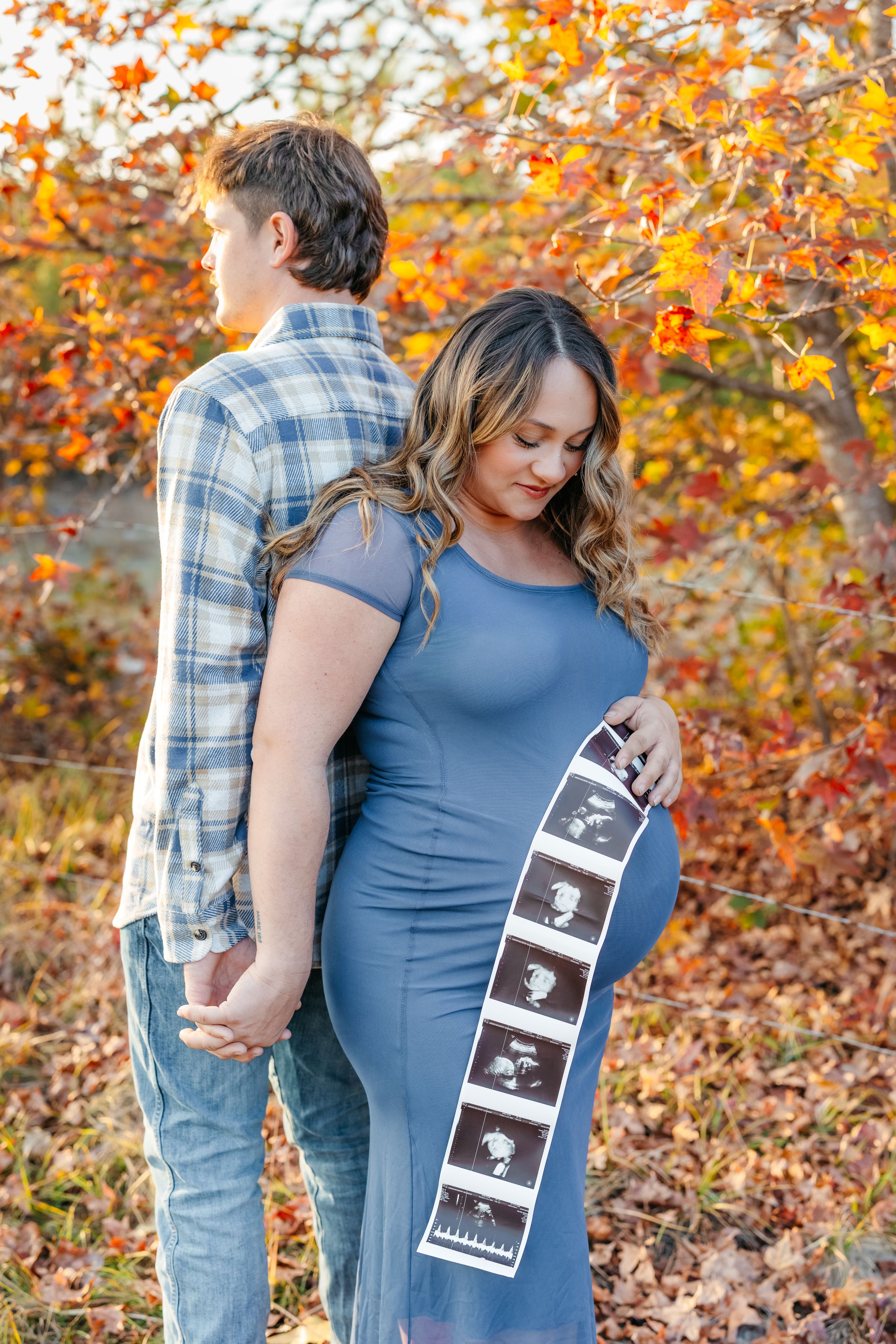 A couple poses in an autumn setting with colorful fall foliage while displaying ultrasound photos on a blue dress.