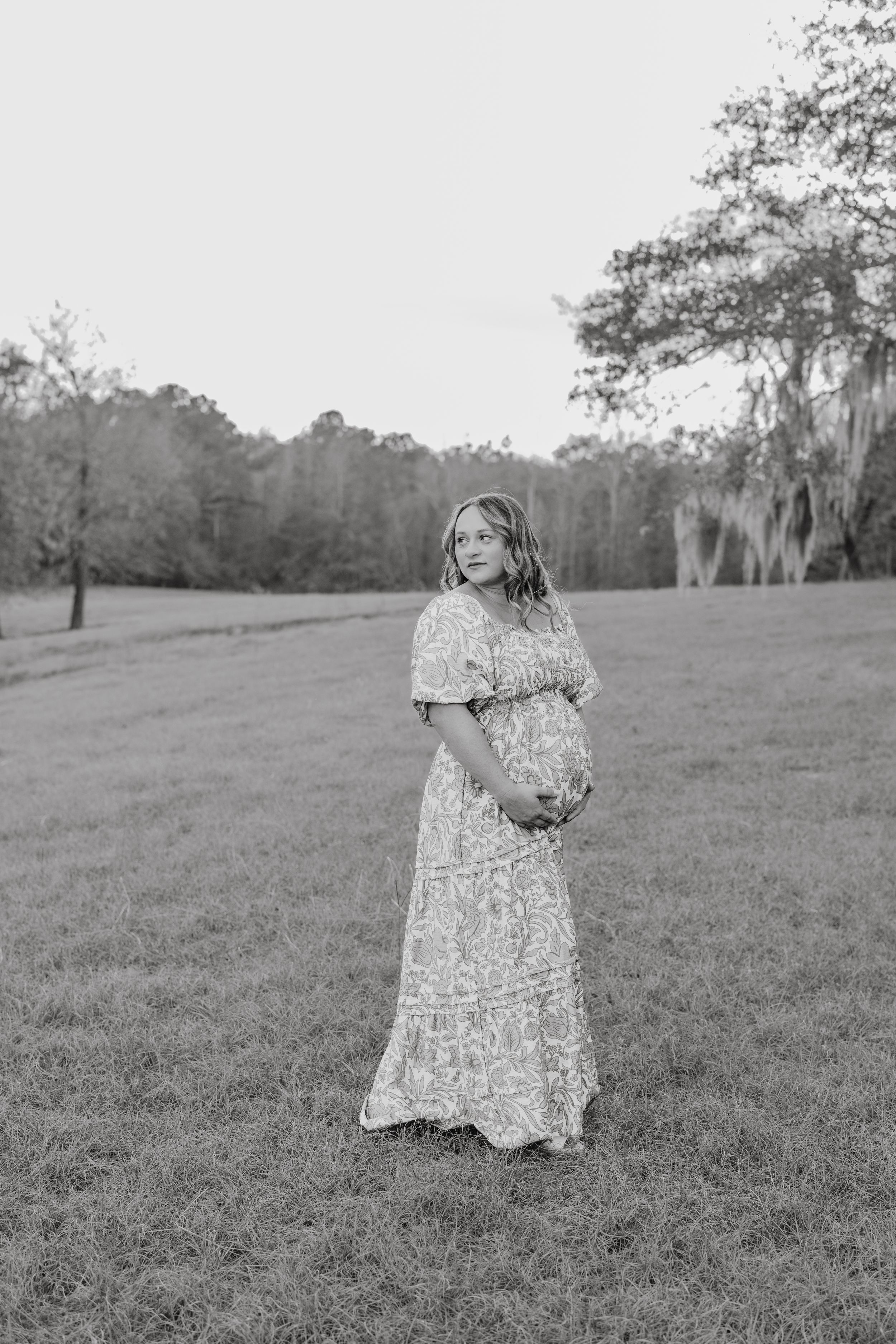 A person in a floral dress stands in an open grassy field with Spanish moss draped trees in the background.