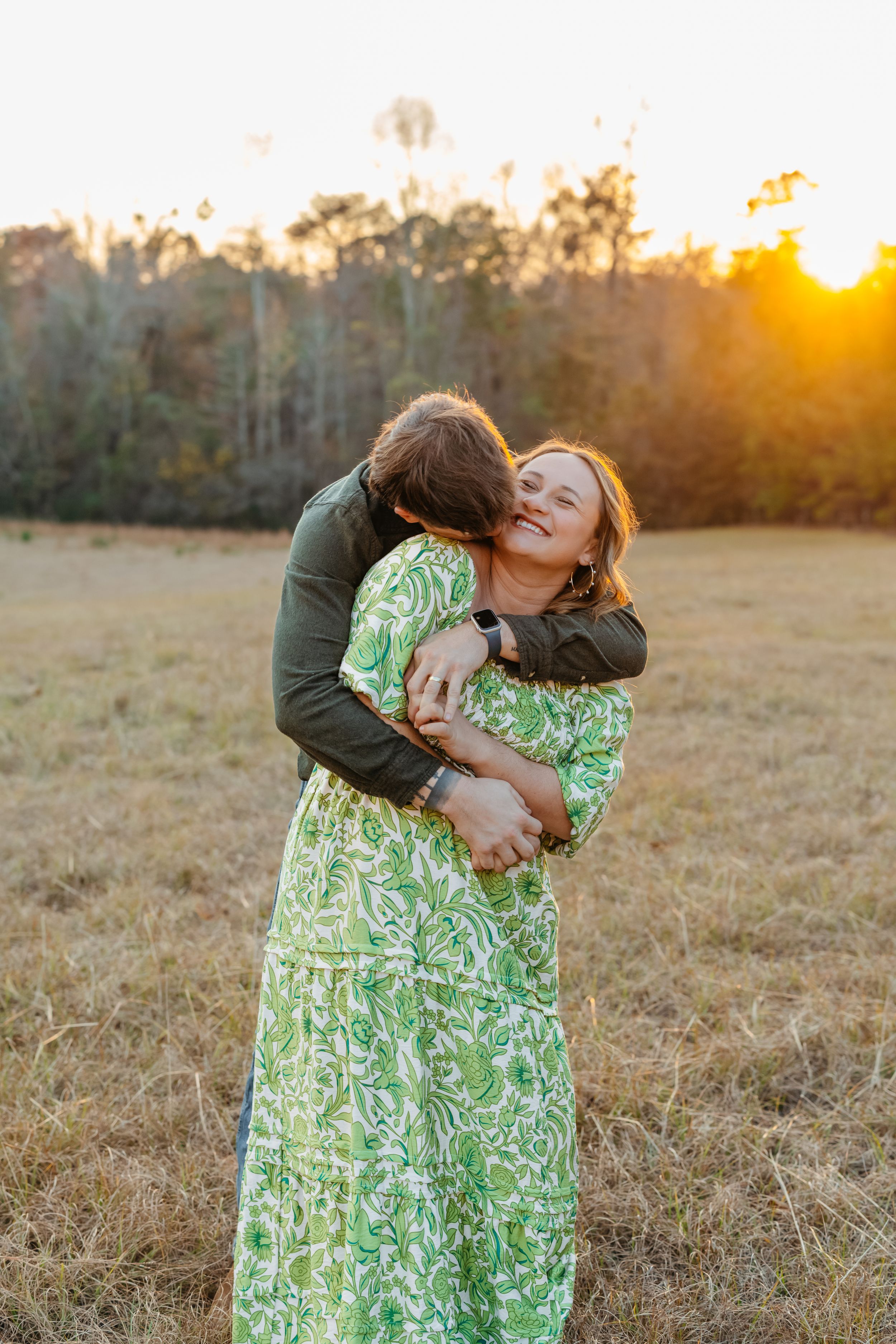 A couple embraces in a golden field at sunset wearing a green floral dress and dark jacket.