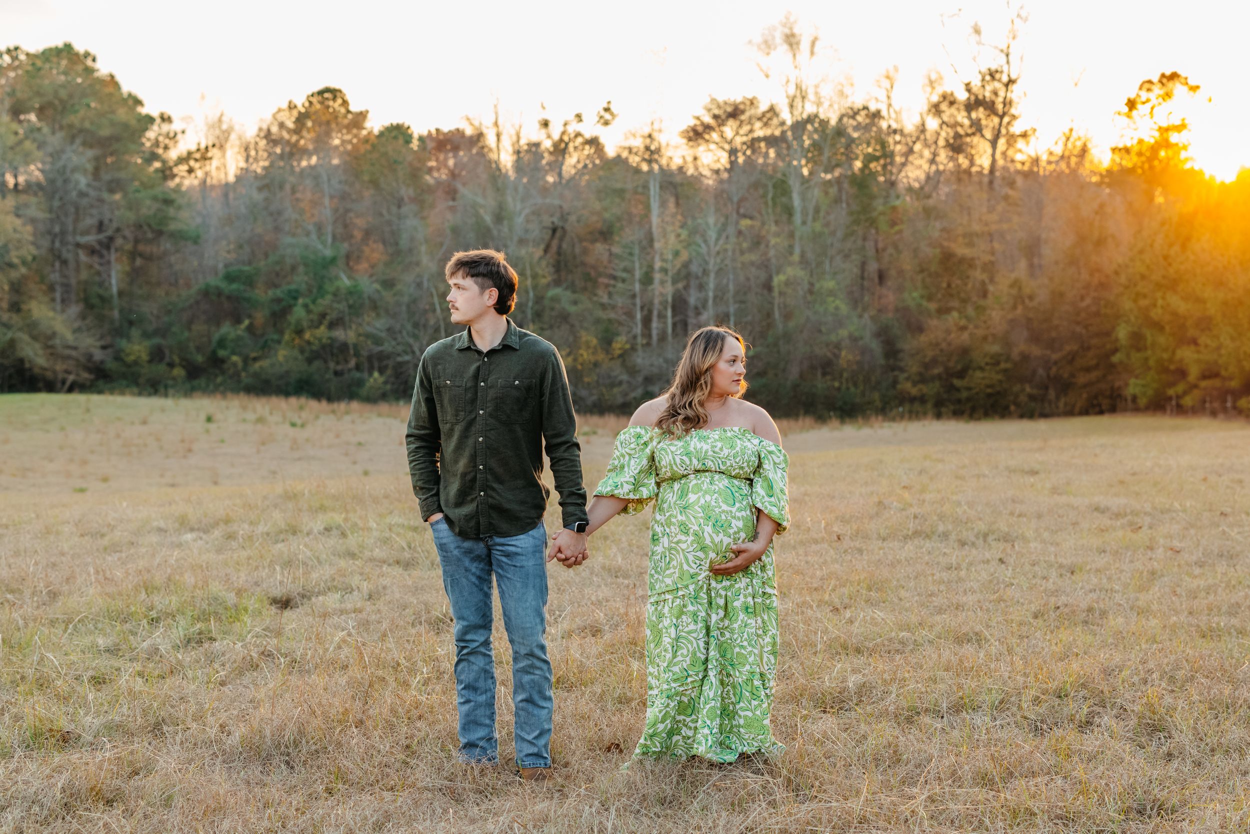 A couple walks hand in hand through a golden field at sunset while wearing casual attire.