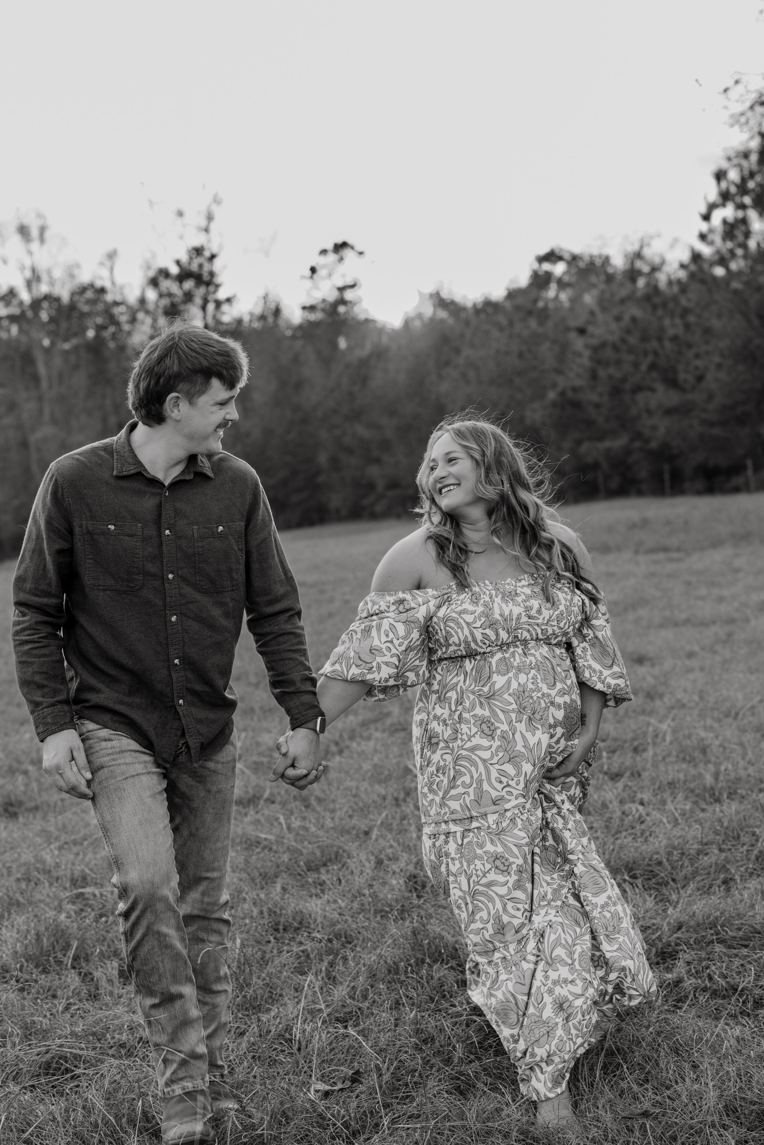 A couple shares playful moments in a grassy field, captured in black and white photography.