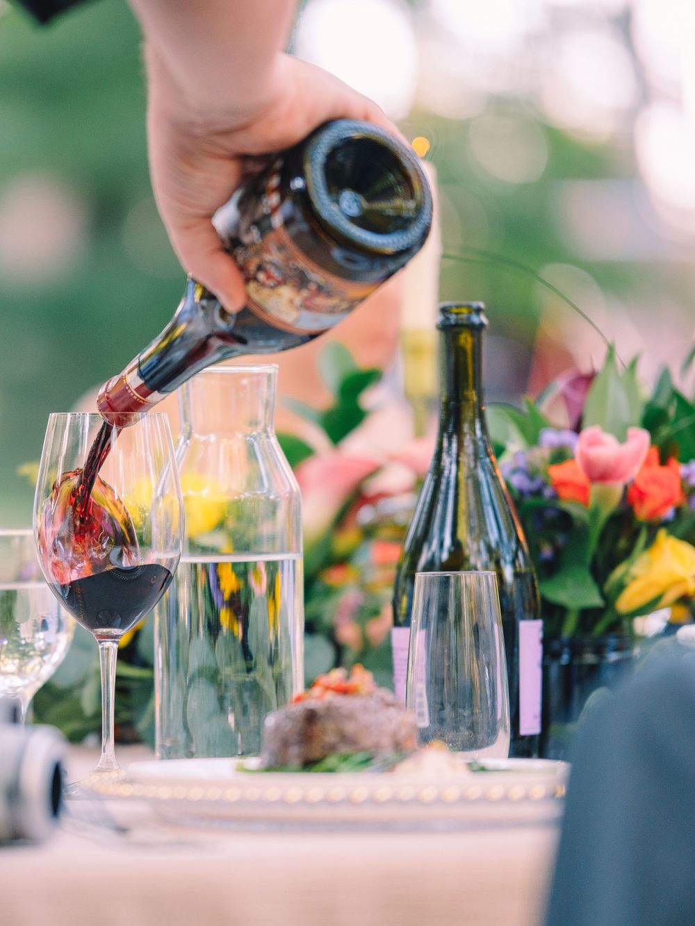 Hand pouring red wine into glass at outdoor table setting with flowers.