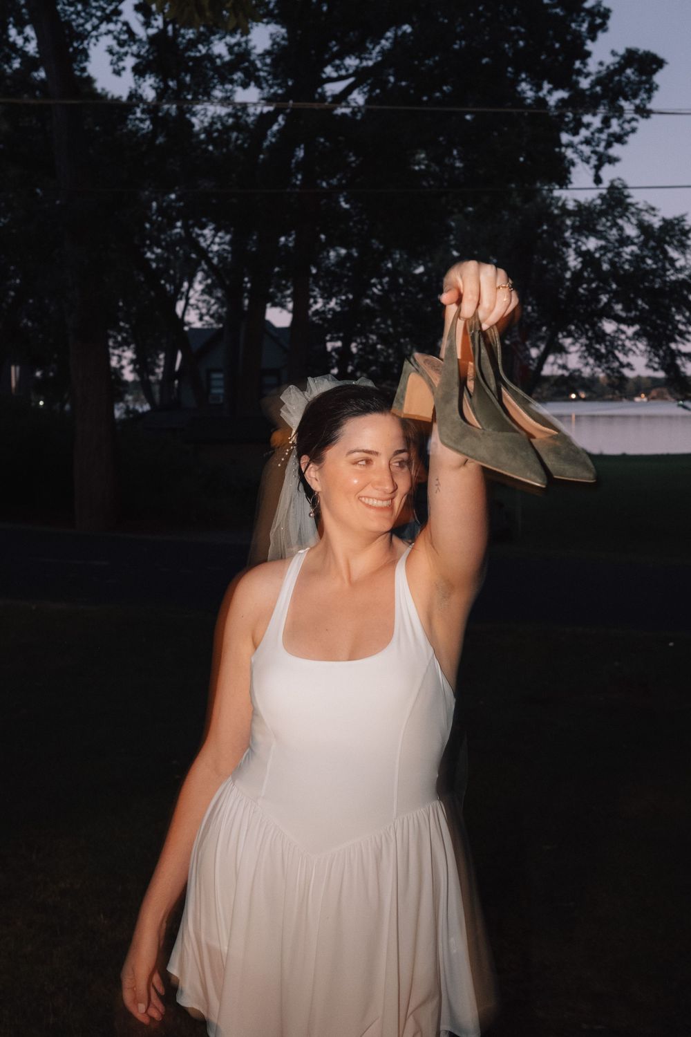 A person in a white dress holds up brown shoes during an outdoor evening celebration.