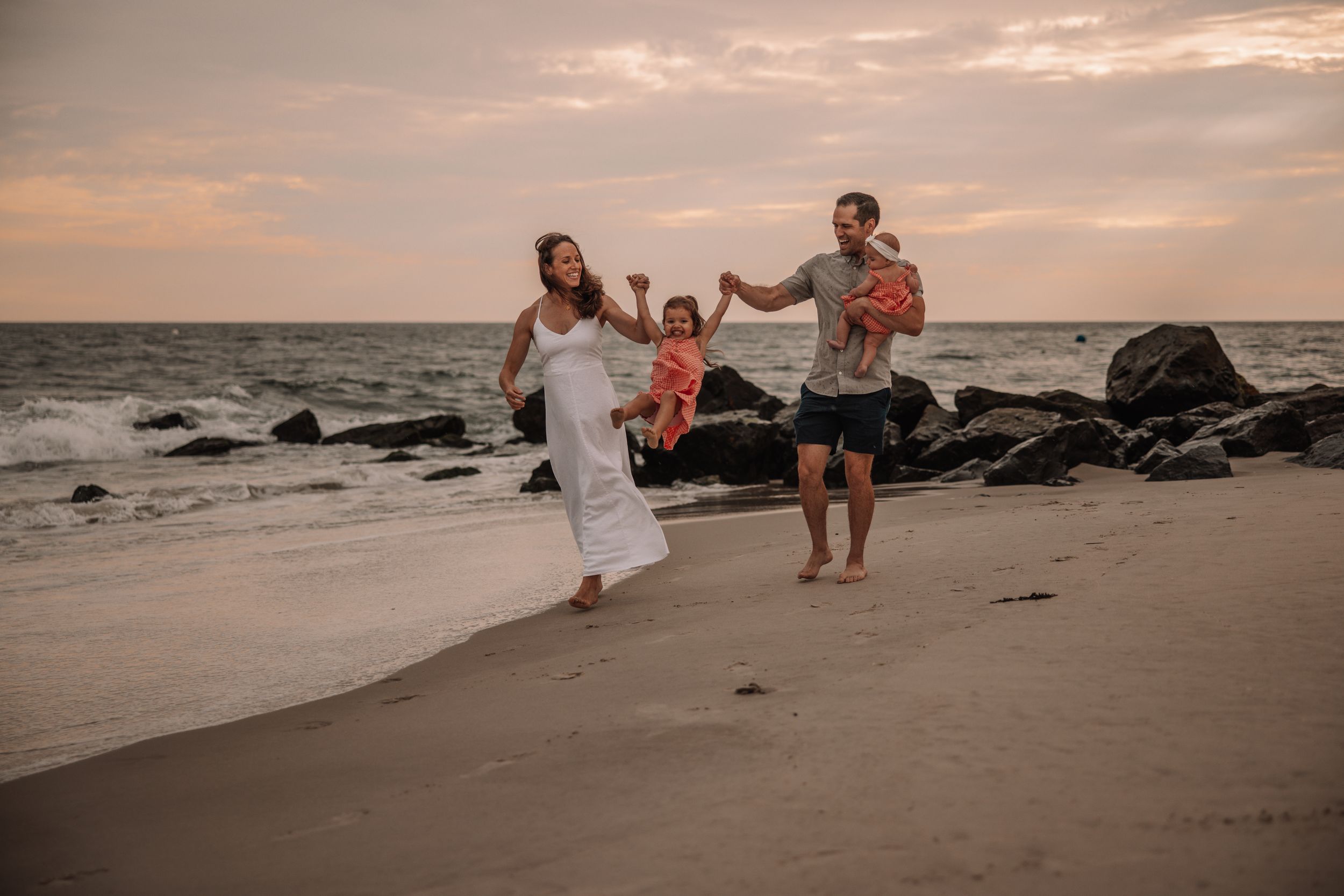 Family of three walking on a sandy beach at sunset, mother in white dress, father holding toddler near rocky shoreline.