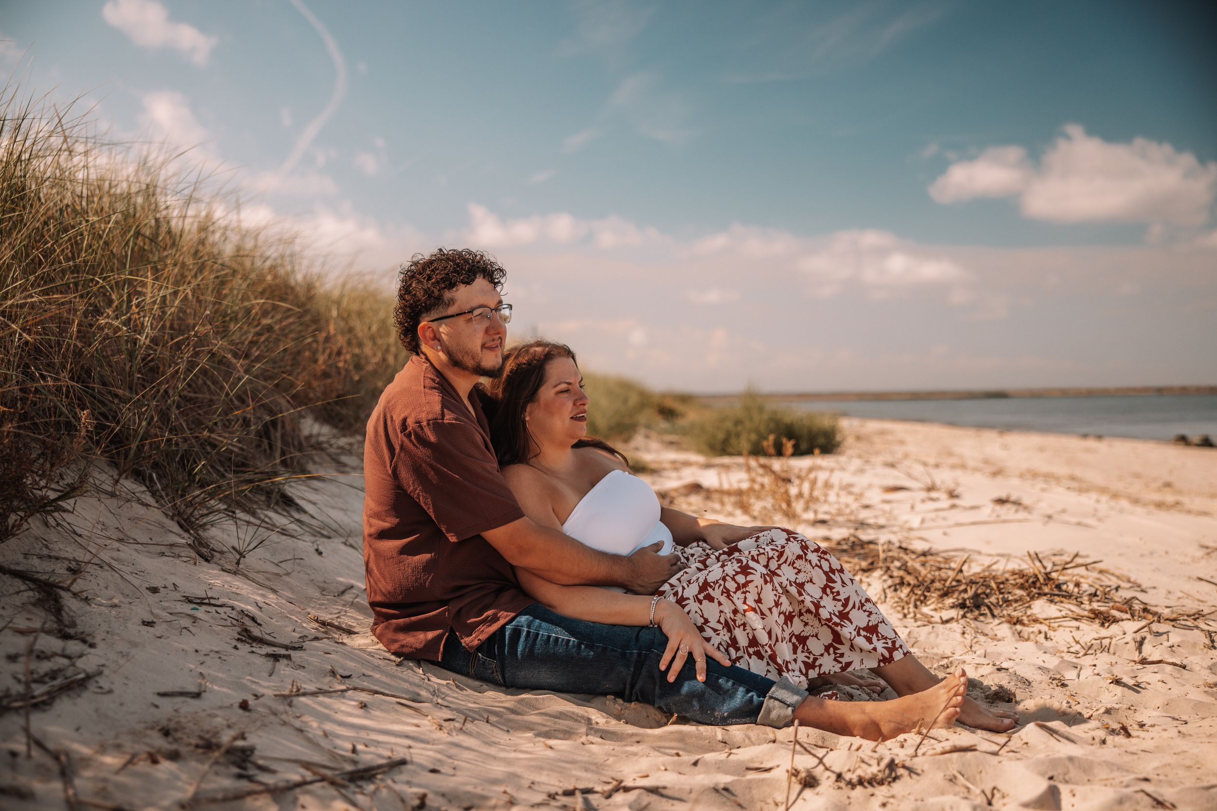 Couple sitting together on sandy beach dunes, embracing and gazing at the ocean under a partly cloudy sky.