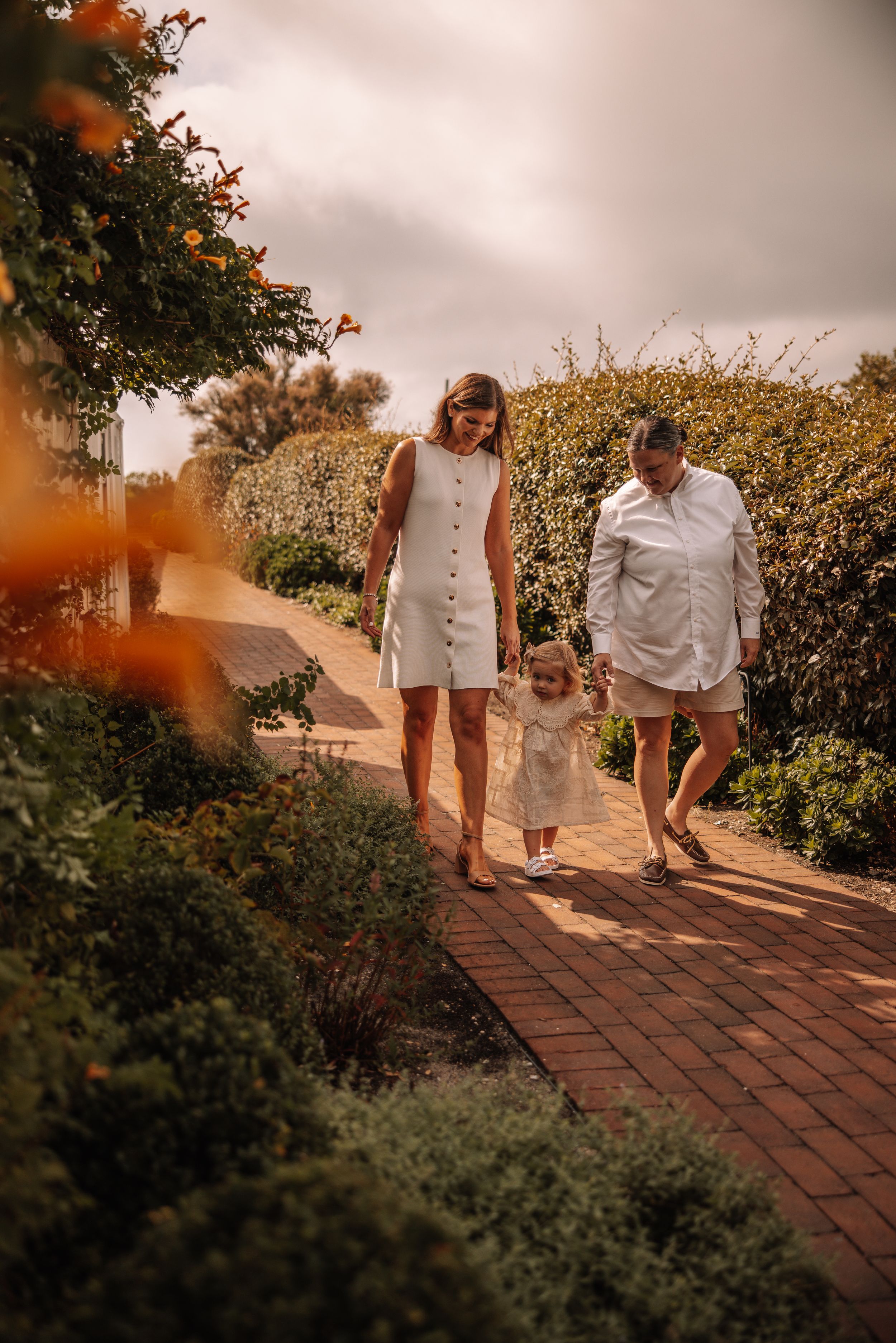 Two women and a toddler walk hand-in-hand along a brick garden path surrounded by lush greenery and orange flowers.