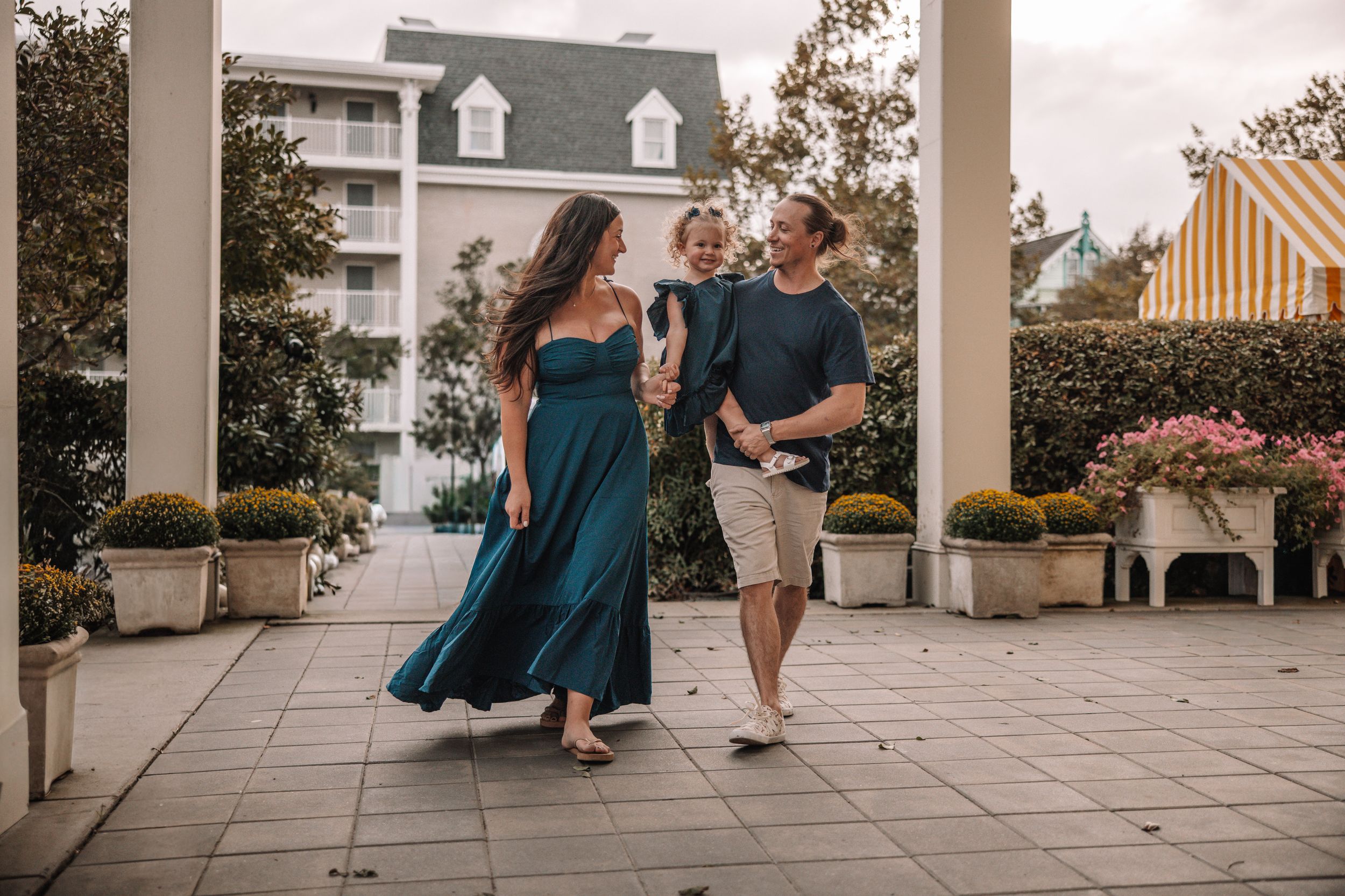 Happy family with toddler strolling on elegant patio, woman in teal gown, man in casual attire, resort backdrop.