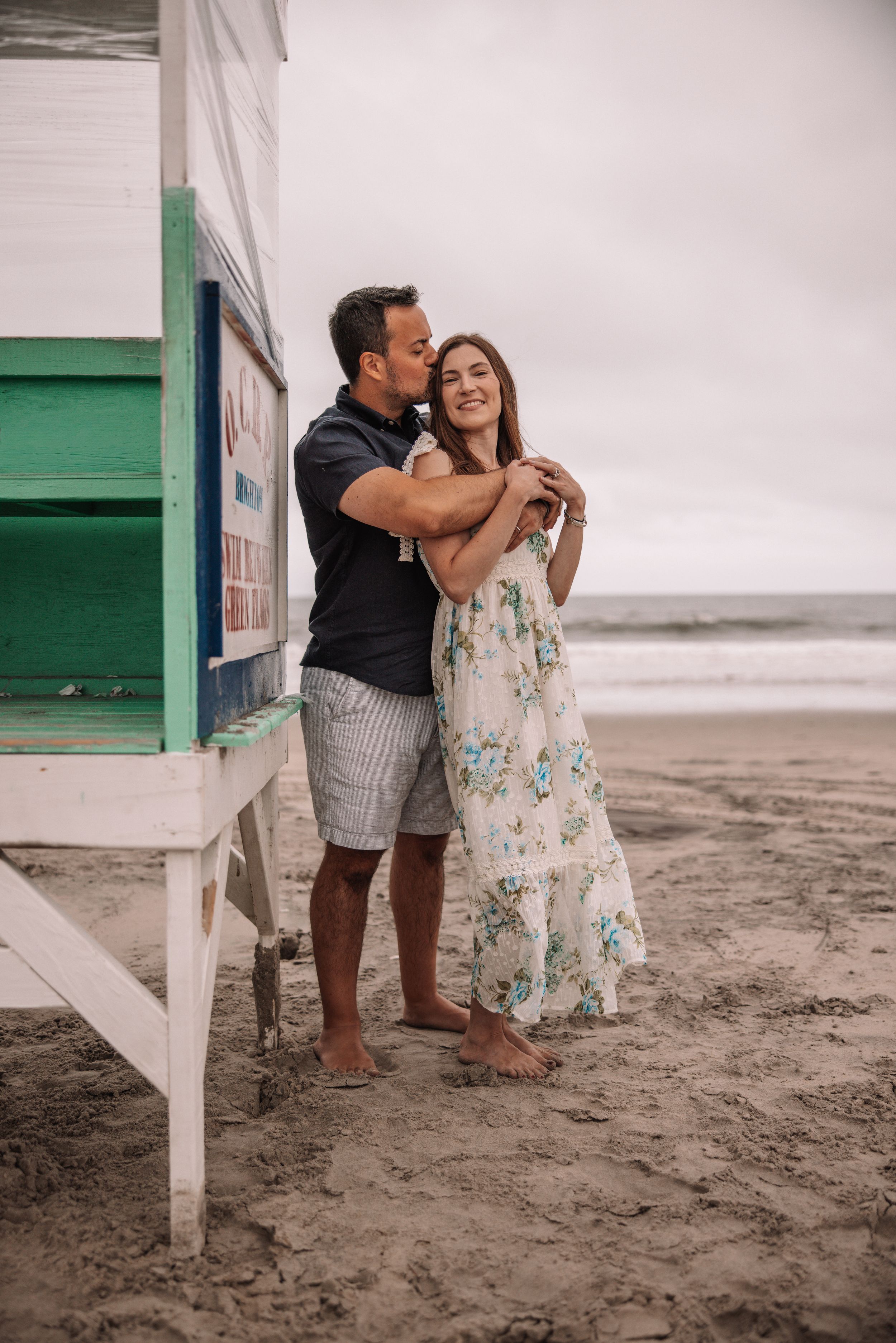 Couple embracing on a sandy beach near a lifeguard stand, woman in floral dress, man in black shirt and gray shorts.