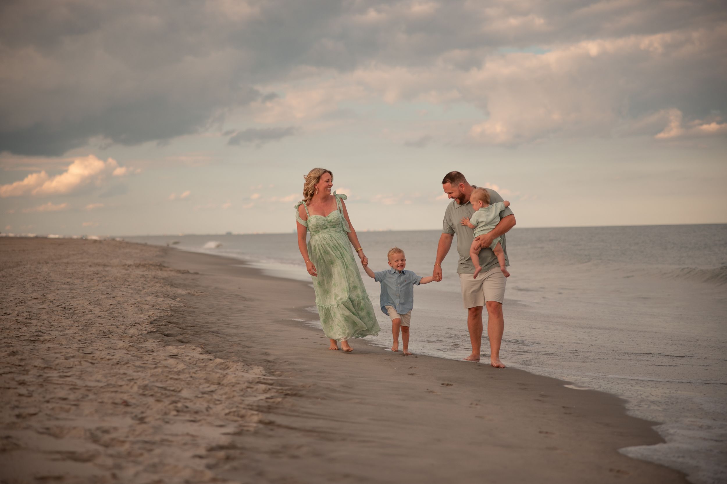 Family of four walking on a beach at sunset, pregnant mother in floral dress, father carrying child.