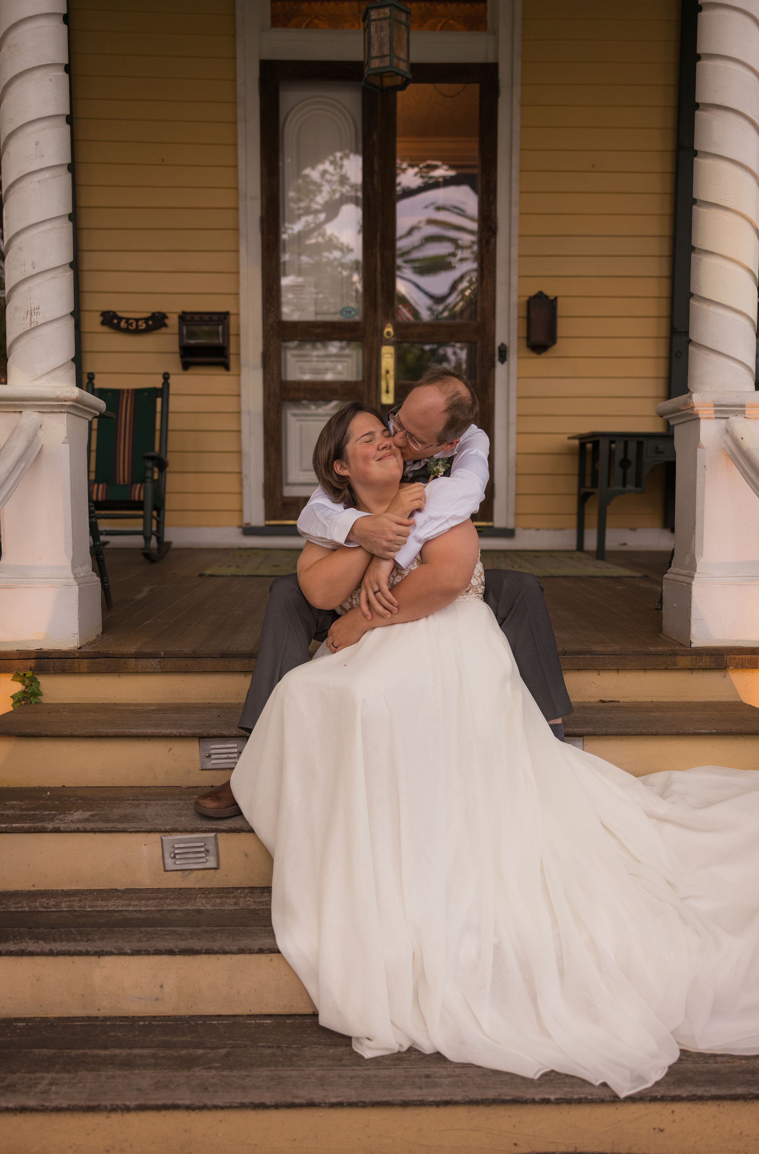 A romantic wedding moment captured on the front porch steps of a yellow house with white columns.