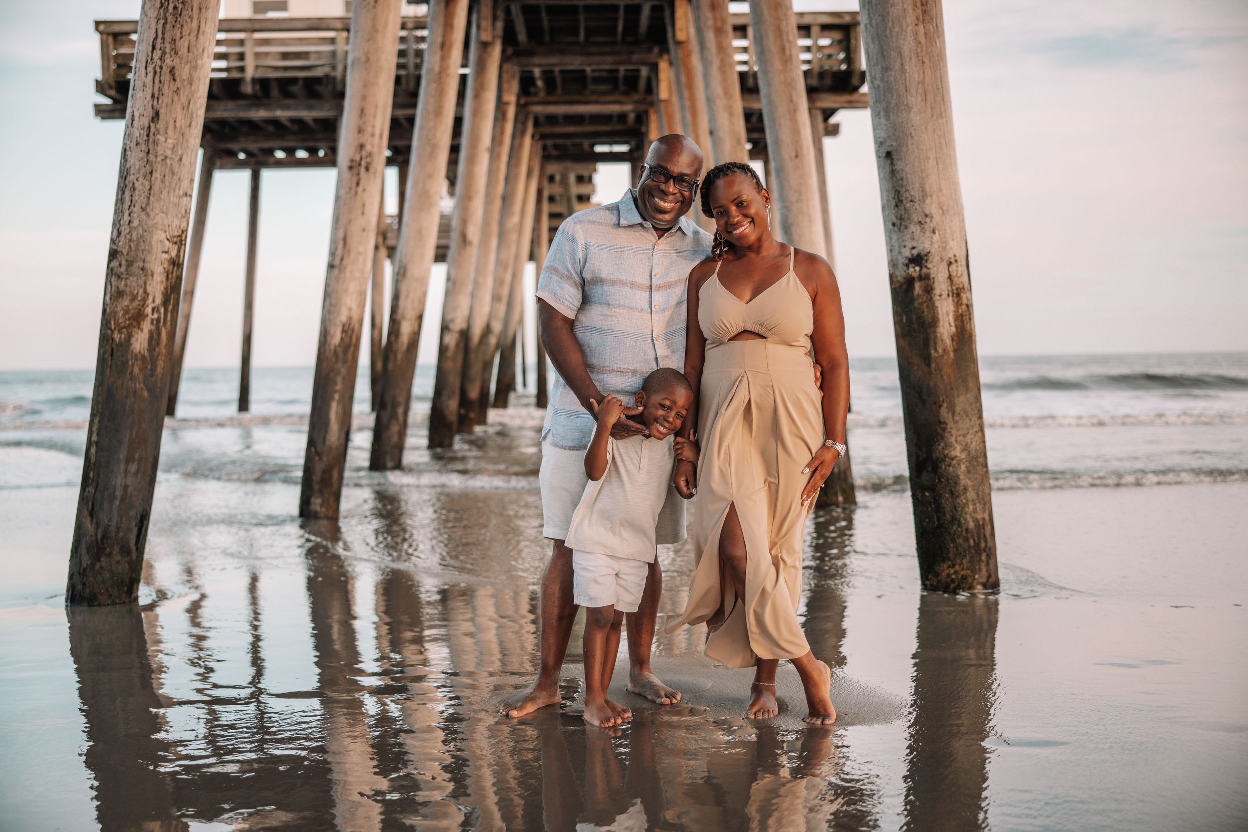 Family of three posing under a beach pier, with a pregnant mother in a beige dress, reflecting on wet sand.
