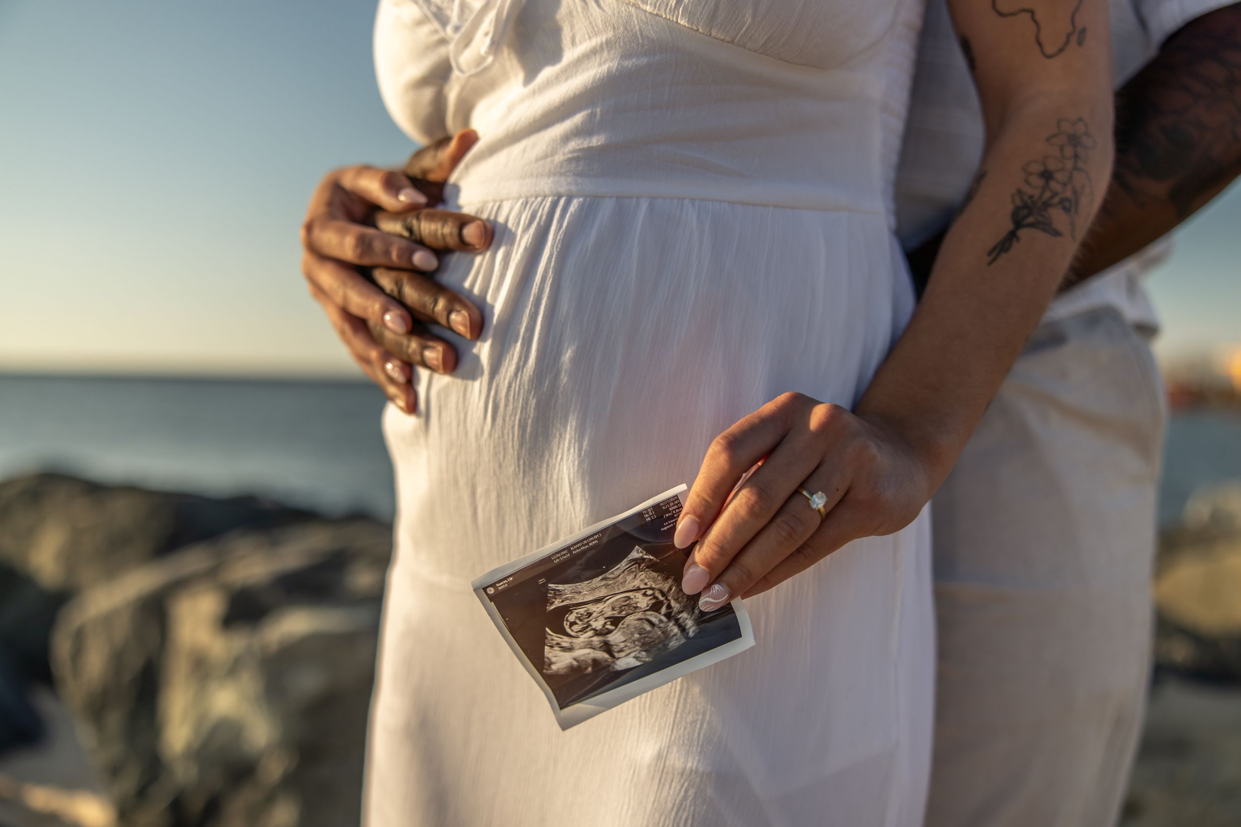 Pregnant couple in white dress holding ultrasound photo by rocky beach at sunset, showing tattooed arms and baby bump.