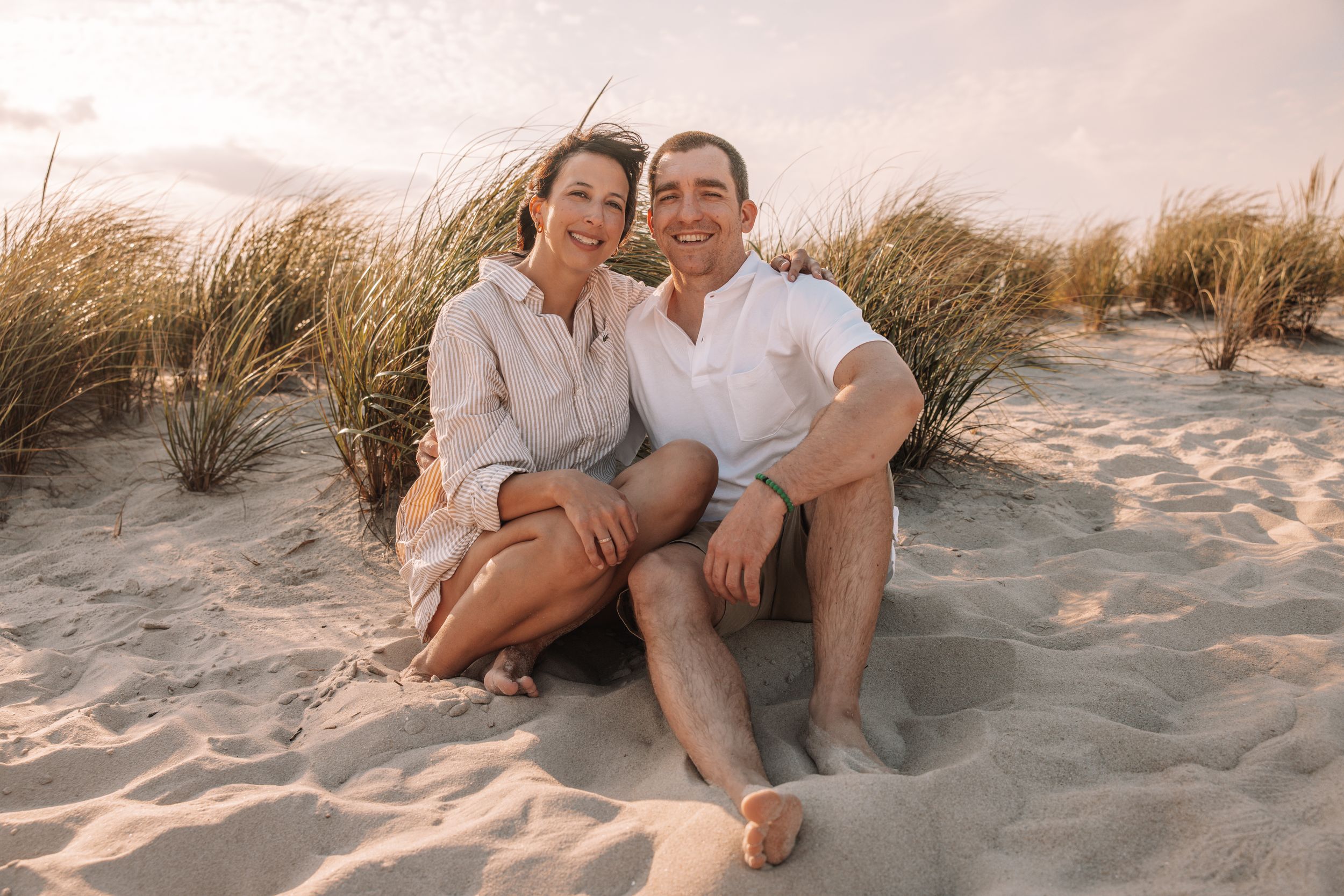 A smiling couple sits together on sandy beach dunes at sunset, dressed casually in white and beige outfits.