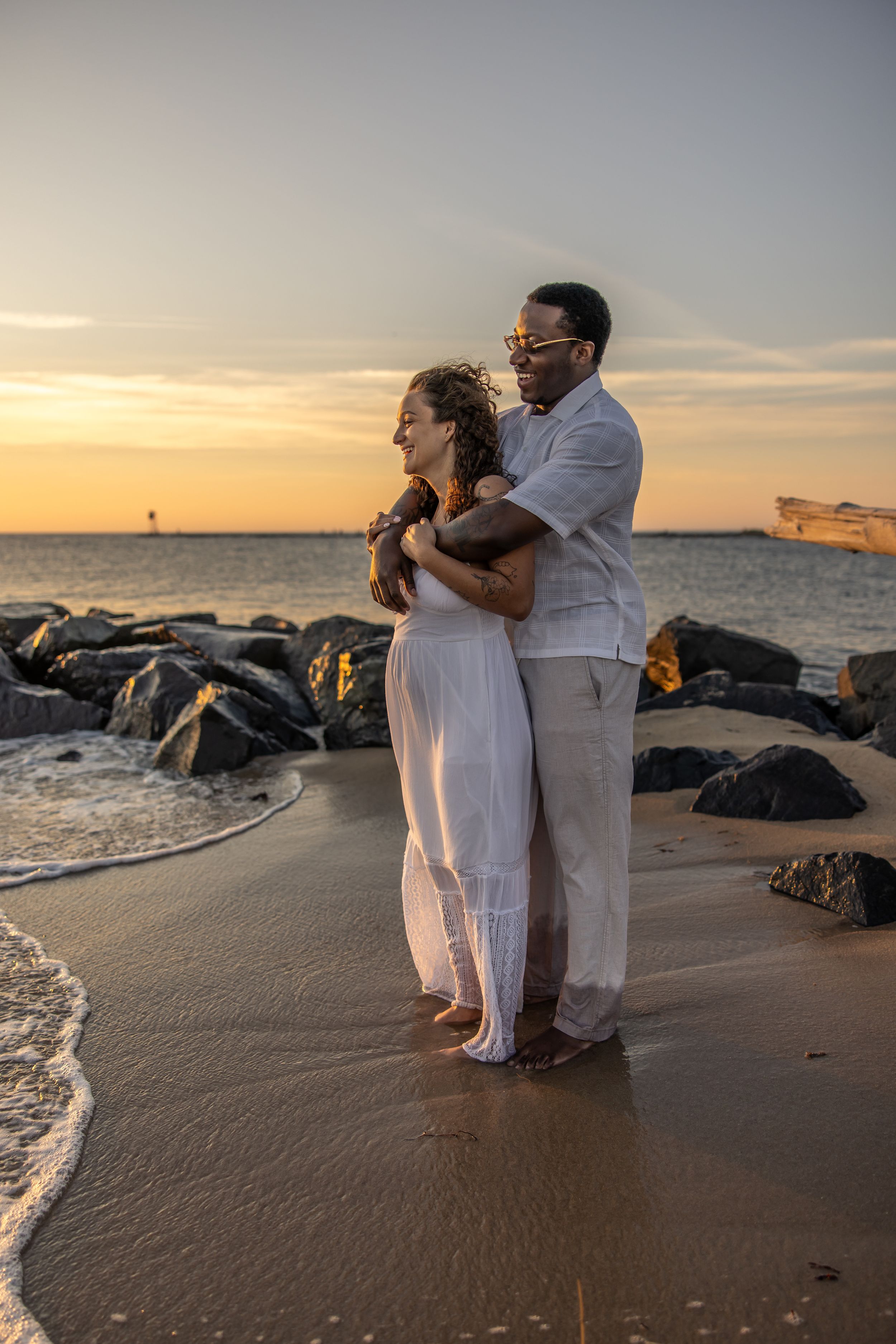 Couple in white embracing on a sandy beach at sunset, with rocks and ocean waves in the background.