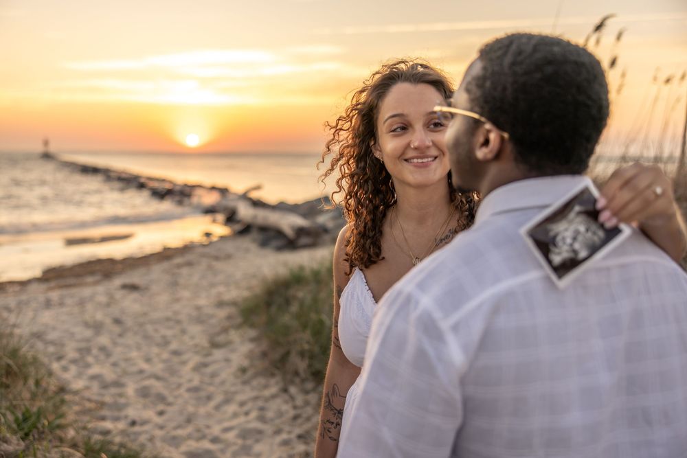 Couple on beach at sunset, woman smiling as man kisses her cheek while holding an ultrasound photo.