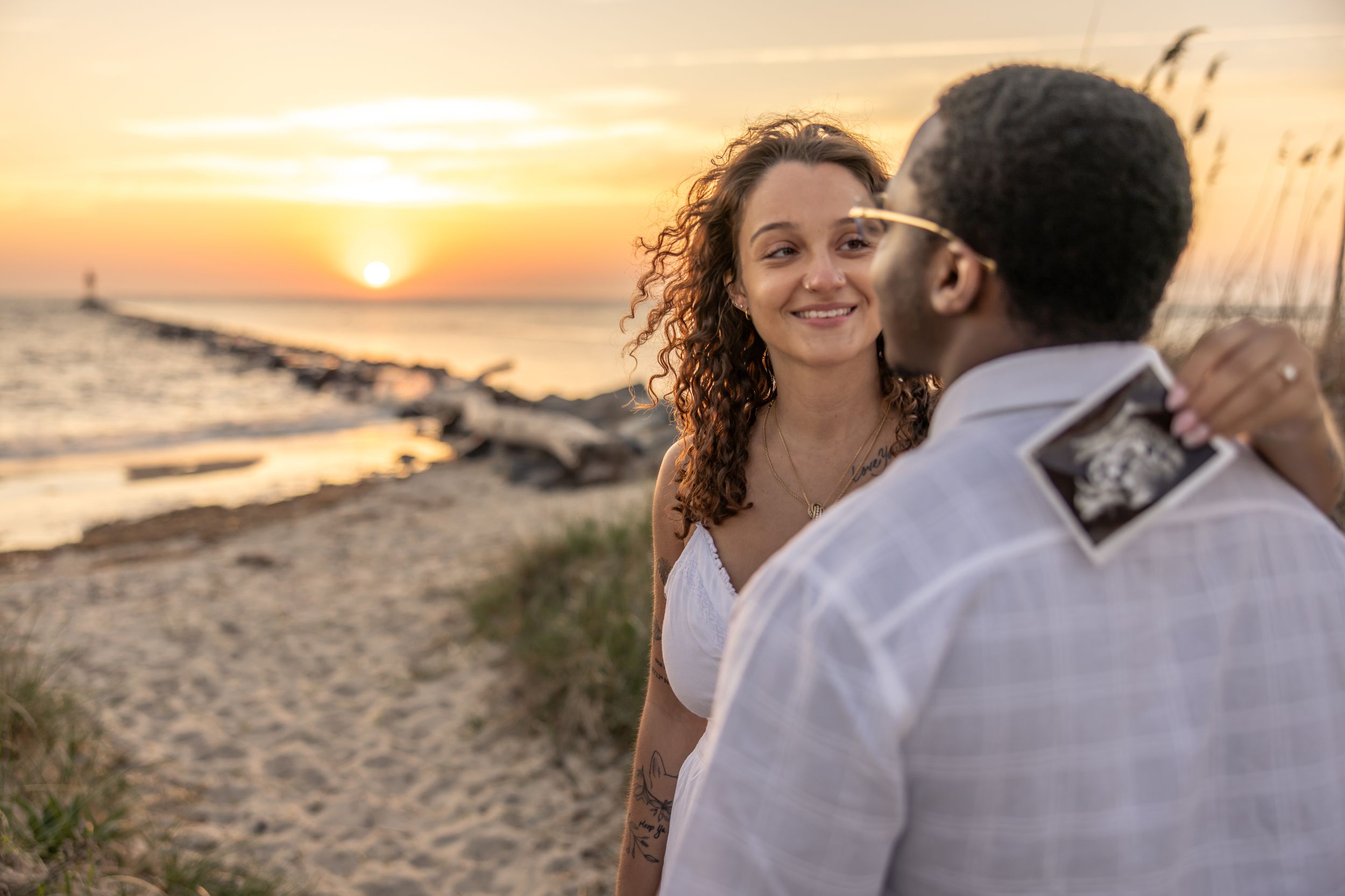 Couple on beach at sunset, woman smiling as man kisses her cheek while holding an ultrasound photo.